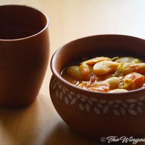 Butter beans curry in an earthen vessel next to an earthen water glass on a brown table.