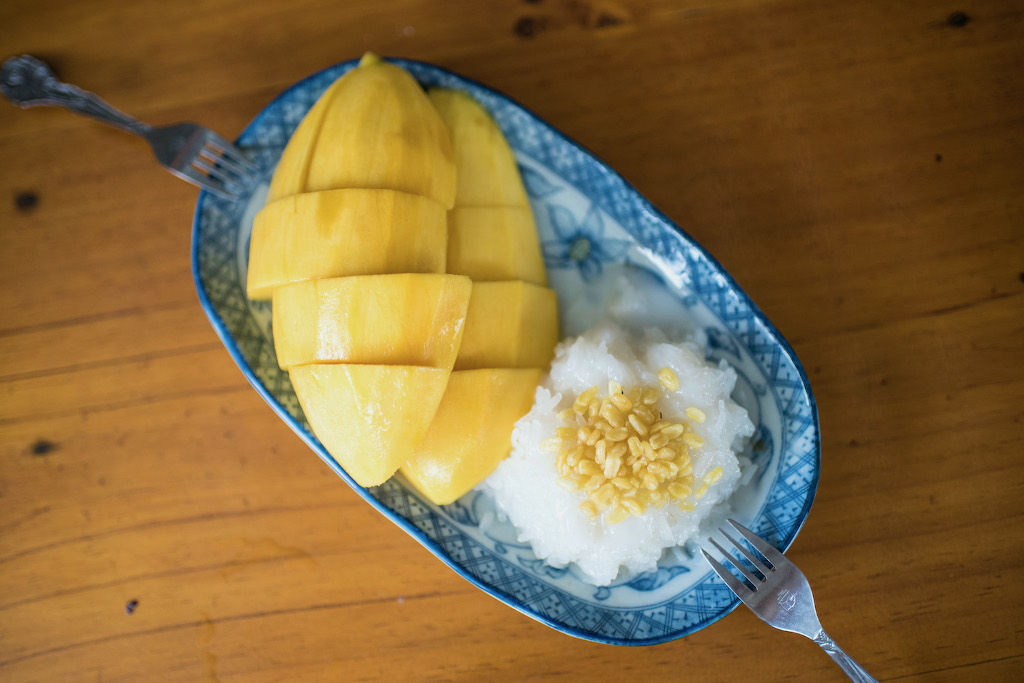 Glutinous rice and fresh mango slices on a blue and white plate with two forks on a brown table. 