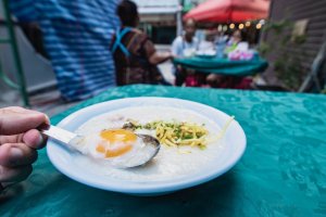 Thai congee topped with an egg in a white bowl on a blue table. Thai congee is a streetfood consisting of rice porridge served with pork or meat that's eaten for breakfast in Thailand. Veg versions are available too.