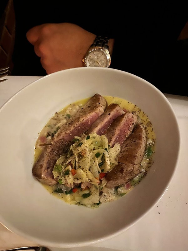 Meat in a white dish at a restaurant with a man's hand with a watch next to it.
