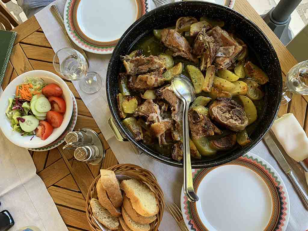 Slow-cooked meat and vegetables in iron pan with bread and salad served on a table.